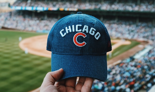 A hand holds a Chicago Cubs baseball cap with the packed stands of Wrigley Field in the background